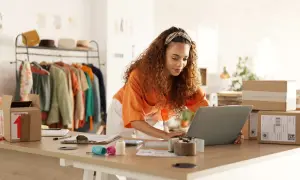 Mujer con cabello rizado trabajando en su laptop para crear una web de venta online. Hay ropa en percheros y cajas de envío listas.