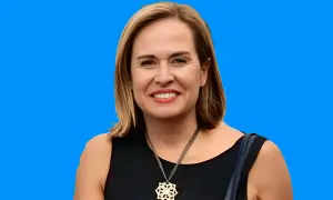 Elena Gómez del Pozuelo sonríe con un collar llamativo, vestida con un elegante traje negro sobre un fondo azul vibrante.
