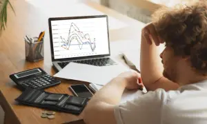 Hombre frustrado con gráfico de trading en pantalla. Calculadora, billetera vacía y monedas sugieren pérdidas.