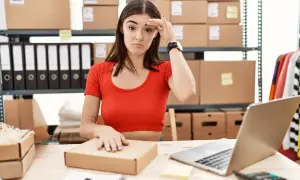 Mujer frustrada frente a un portátil, rodeada de cajas y material de oficina, lidiando con la gestión de stock.
