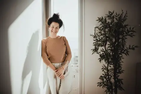 Mujer sonriente apoya en pared, con planta y luz. Sus youtoproject opiniones son positivas.