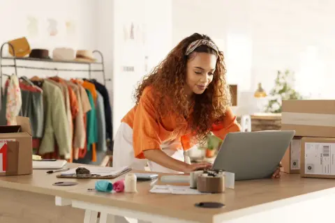 Mujer con cabello rizado trabajando en su laptop para crear una web de venta online. Hay ropa en percheros y cajas de envío listas.