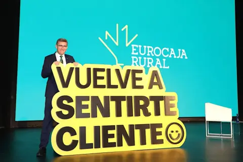 Hombre sonriente con traje sostiene cartel amarillo con mensaje "Vuelve a sentirte cliente" y logo de Eurocaja Rural.