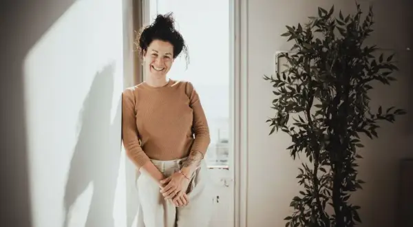 Mujer sonriente apoya en pared, con planta y luz. Sus youtoproject opiniones son positivas.