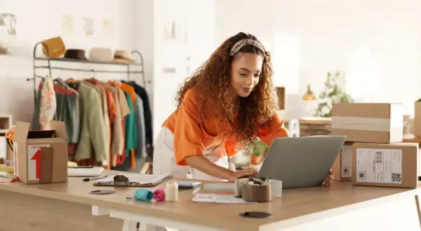 Mujer con cabello rizado trabajando en su laptop para crear una web de venta online. Hay ropa en percheros y cajas de envío listas.