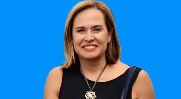 Elena Gómez del Pozuelo sonríe con un collar llamativo, vestida con un elegante traje negro sobre un fondo azul vibrante.