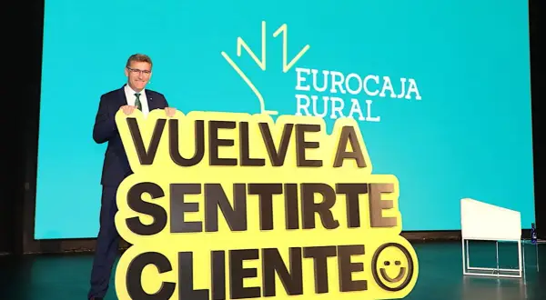 Hombre sonriente con traje sostiene cartel amarillo con mensaje "Vuelve a sentirte cliente" y logo de Eurocaja Rural.