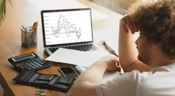 Hombre frustrado con gráfico de trading en pantalla. Calculadora, billetera vacía y monedas sugieren pérdidas.