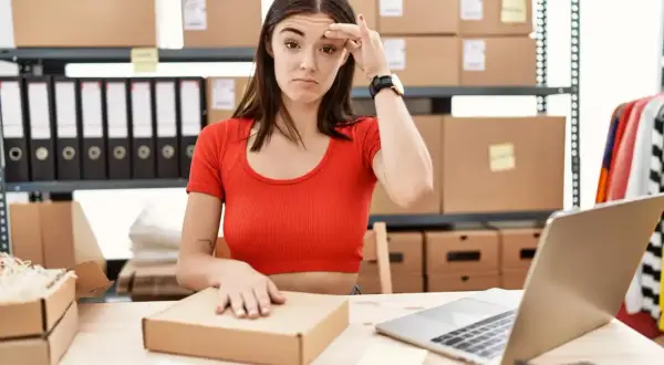 Mujer frustrada frente a un portátil, rodeada de cajas y material de oficina, lidiando con la gestión de stock.