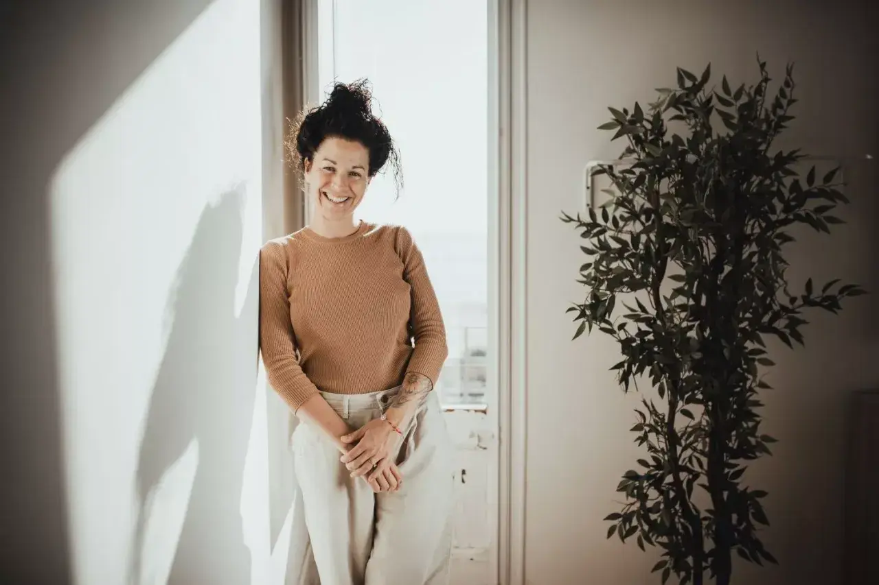 Mujer sonriente apoya en pared, con planta y luz. Sus youtoproject opiniones son positivas.