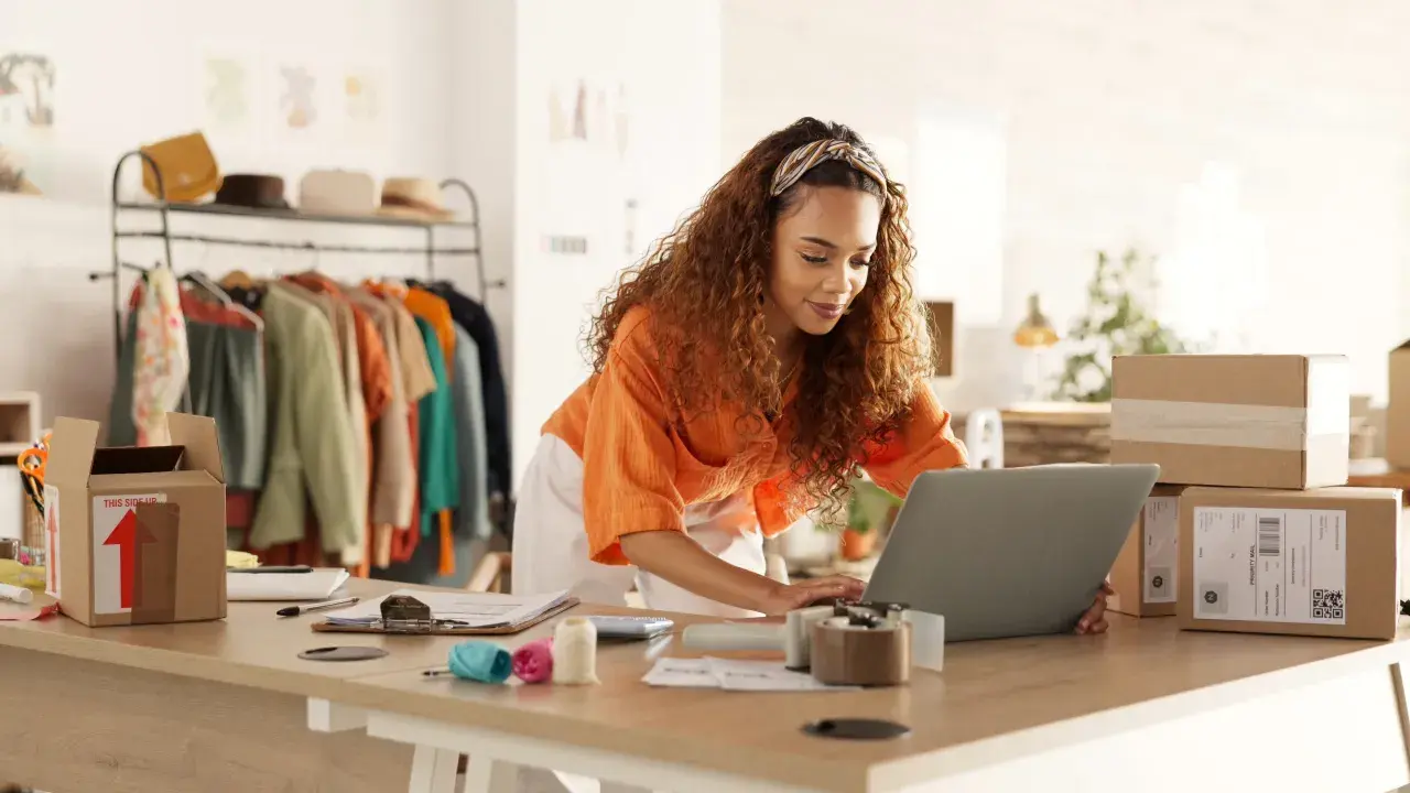 Mujer con cabello rizado trabajando en su laptop para crear una web de venta online. Hay ropa en percheros y cajas de envío listas.