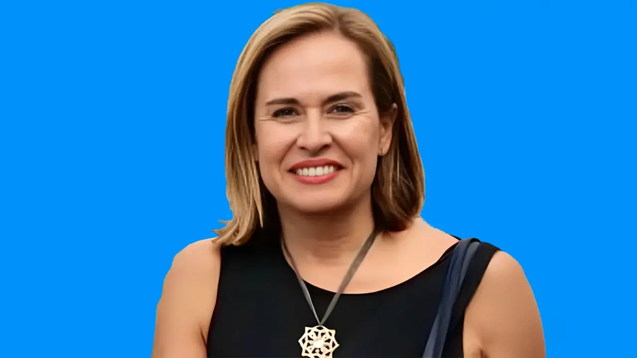 Elena Gómez del Pozuelo sonríe con un collar llamativo, vestida con un elegante traje negro sobre un fondo azul vibrante.