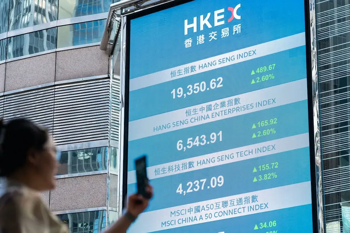 Una mujer mira un teléfono móvil frente a una pantalla que muestra el índice Hang Seng y otros datos bursátiles.