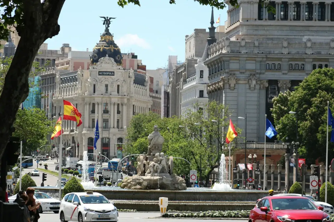 Edificio Metropolis en Madrid, con banderas espa&ntilde;olas ondeando. El tr&aacute;fico fluye, sugiriendo la actividad de negocios millonarios en esta vibrante ciudad.