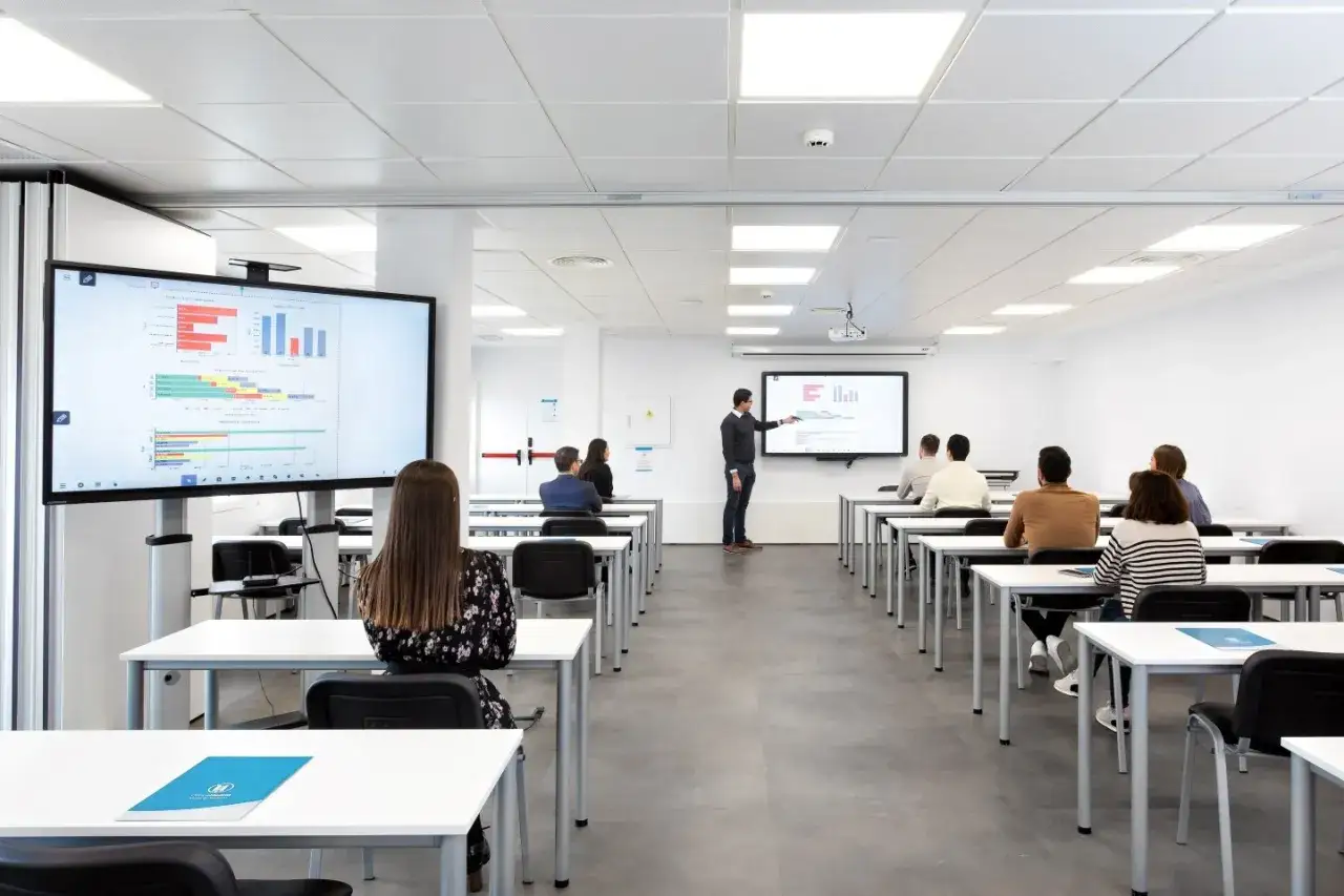 Un profesor explica gr&aacute;ficos en una pantalla grande a estudiantes sentados en un aula moderna, ideal para montar una academia.