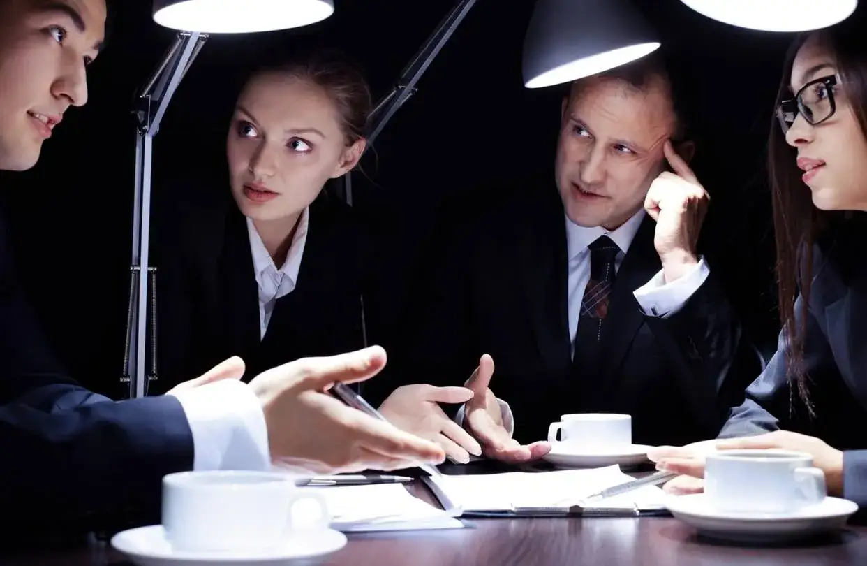 Cuatro personas en trajes negros participan en entrevistas a emprendedores, bajo la luz de l&aacute;mparas de escritorio.