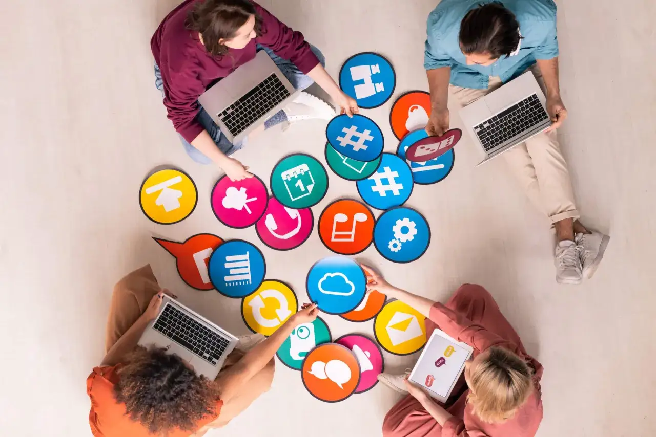Un grupo de personas colabora en el suelo, rodeado de iconos de aplicaciones de productividad como nube, calendario y m&uacute;sica.