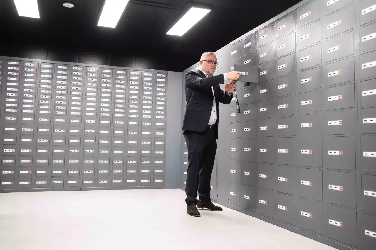 Hombre con gafas abre una caja de seguridad en un banco. La sala está llena de filas de casilleros.