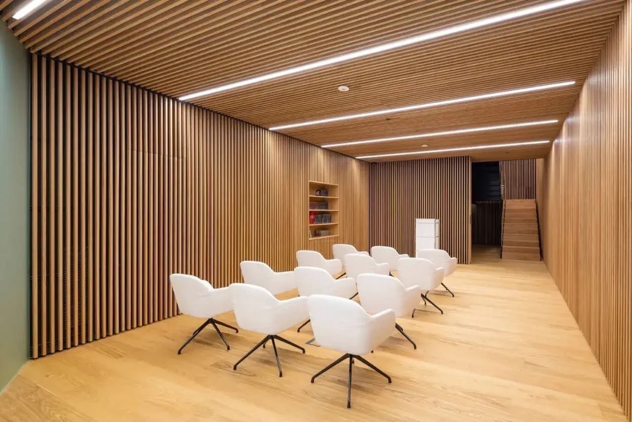 Sala de espera moderna con paredes y techo de madera, sillas blancas y una escalera al fondo. Un espacio que parece una caja de arquitectos.