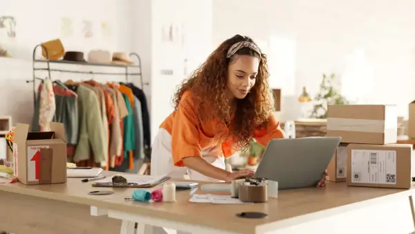 Mujer con cabello rizado trabajando en su laptop para crear una web de venta online. Hay ropa en percheros y cajas de envío listas.