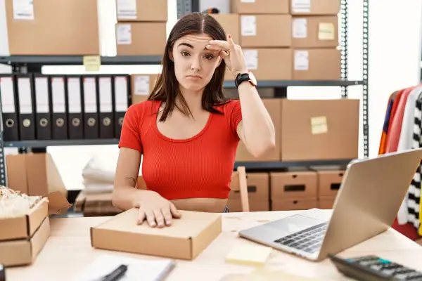 Mujer frustrada revisando el inventario online, rodeada de cajas y carpetas.