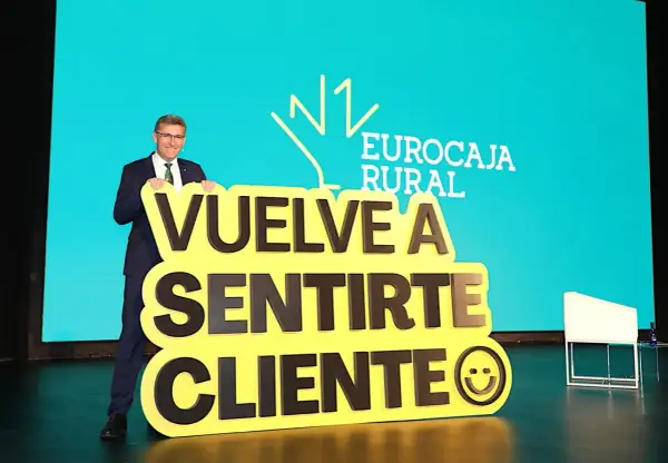 Hombre sonriente con traje sostiene cartel amarillo con mensaje "Vuelve a sentirte cliente" y logo de Eurocaja Rural.