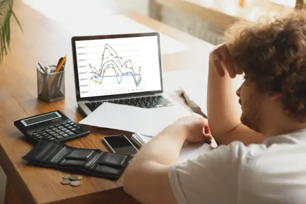 Hombre frustrado con gráfico de trading en pantalla. Calculadora, billetera vacía y monedas sugieren pérdidas.